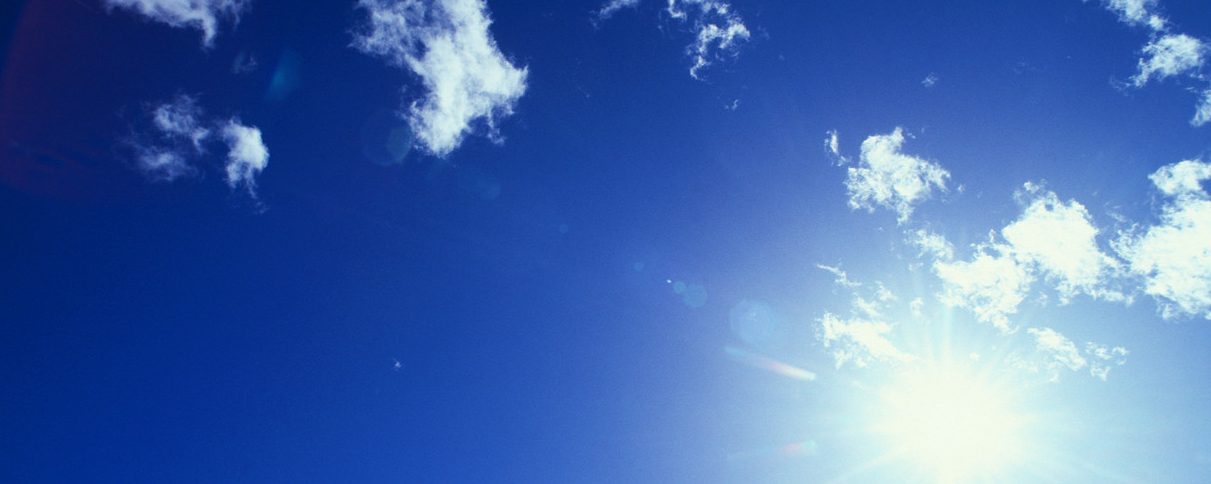 Hellblauer Himmel mit einigen weißen Wolken und der Sonne im Vordergrund.